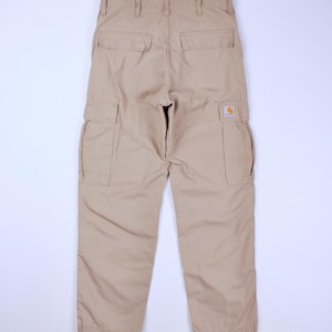 Pode incluir: Cal&ccedil;as cargo bege com v&aacute;rios bolsos e um patch com o logotipo da Carhartt. As cal&ccedil;as t&ecirc;m um ajuste descontra&iacute;do, com cord&otilde;es nos tornozelos. A vista traseira mostra os bolsos e os detalhes das costuras. Um estilo cl&aacute;ssico de roupa de trabalho.