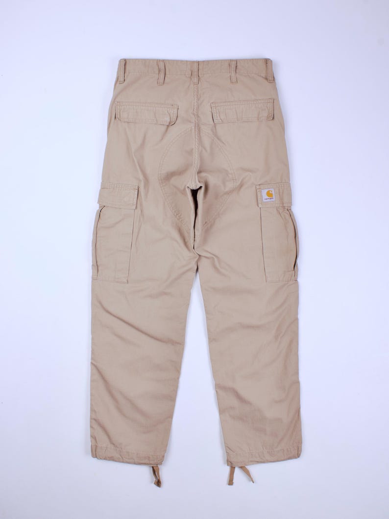 Pode incluir: Cal&ccedil;as cargo bege com v&aacute;rios bolsos e um patch com o logotipo da Carhartt. As cal&ccedil;as t&ecirc;m um ajuste descontra&iacute;do, com cord&otilde;es nos tornozelos. A vista traseira mostra os bolsos e os detalhes das costuras. Um estilo cl&aacute;ssico de roupa de trabalho.