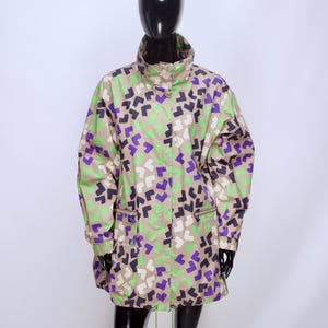 May include: A beige jacket featuring a repeating geometric pattern of small, angled shapes in purple, lime green, and white. The jacket includes a high collar, long sleeves with elastic cuffs, a front zipper closure, and two lower pockets with flaps.