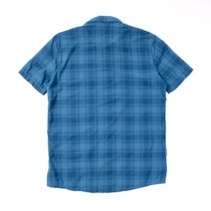 Puede incluir: Una camisa de manga corta con botones en un tono azul medio con un patr&oacute;n de cuadros azul m&aacute;s oscuro. La camisa tiene un cuello cl&aacute;sico, un dobladillo recto y una peque&ntilde;a etiqueta rectangular de marca en la costura lateral inferior.
