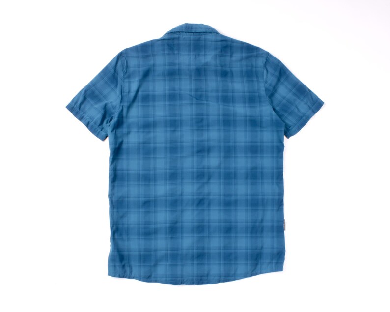 Puede incluir: Una camisa de manga corta con botones en un tono azul medio con un patr&oacute;n de cuadros azul m&aacute;s oscuro. La camisa tiene un cuello cl&aacute;sico, un dobladillo recto y una peque&ntilde;a etiqueta rectangular de marca en la costura lateral inferior.