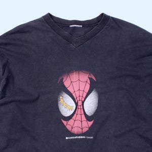 May include: A black long-sleeve V-neck shirt featuring a large graphic of Spider-Man's mask. The mask is red with a white eye design and a web pattern. The word "Spider-Man" is in yellow on the eye. The shirt has the text "SENSATIONAL" at the bottom.