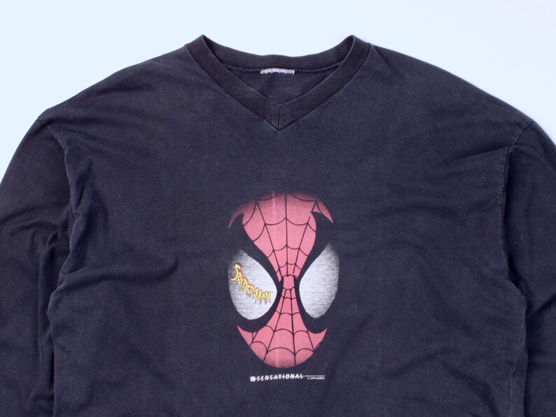 May include: A black long-sleeve V-neck shirt featuring a large graphic of Spider-Man's mask. The mask is red with a white eye design and a web pattern. The word "Spider-Man" is in yellow on the eye. The shirt has the text "SENSATIONAL" at the bottom.
