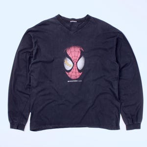 May include: Black long-sleeve shirt with a V-neck. The shirt features a graphic of Spider-Man's mask in red and white, with yellow eye details. The shirt is laid flat on a white surface.