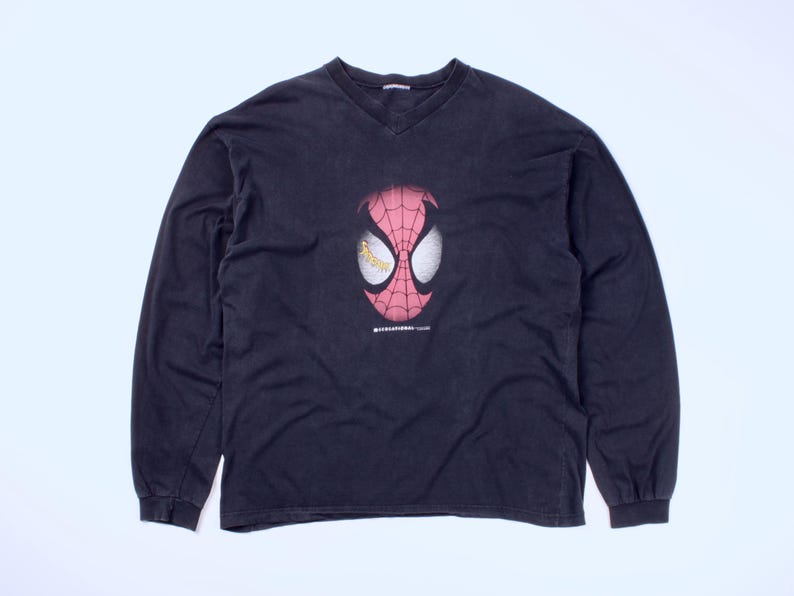May include: Black long-sleeve shirt with a V-neck. The shirt features a graphic of Spider-Man's mask in red and white, with yellow eye details. The shirt is laid flat on a white surface.