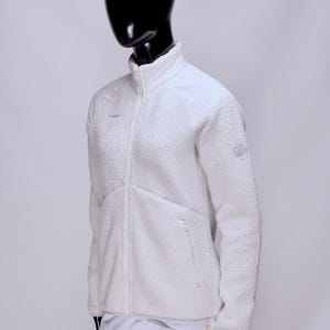 May include: A white fleece jacket with a full-length zipper, side pockets, and a high collar. The jacket is displayed on a mannequin. The Mammut logo is visible on the chest and sleeve. The jacket is made of a soft, textured material.