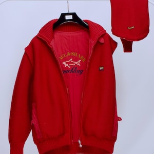 May include: A red zip-up jacket and matching scarf set. The jacket features a ribbed collar, a front zipper, and a small logo patch. The inner lining of the jacket has a logo with the text "MCL & SHARK yachting".