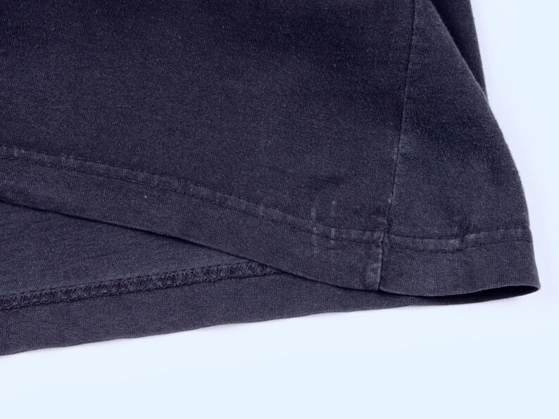 May include: Close-up of a dark gray t-shirt hem. The fabric is folded over, revealing the stitched seam and the edge of the shirt. The material appears to be soft and slightly worn.
