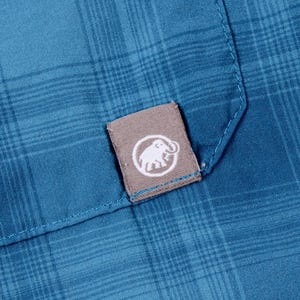 Puede incluir: Una camisa de cuadros azul con una peque&ntilde;a etiqueta de tela gris rectangular cosida en el bolsillo. La etiqueta muestra un logotipo circular blanco con una ilustraci&oacute;n gr&aacute;fica de un mamut. La tela tiene un patr&oacute;n visible de l&iacute;neas cruzadas.