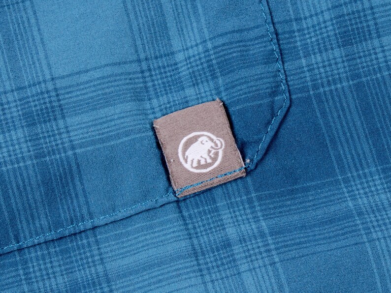 Puede incluir: Una camisa de cuadros azul con una peque&ntilde;a etiqueta de tela gris rectangular cosida en el bolsillo. La etiqueta muestra un logotipo circular blanco con una ilustraci&oacute;n gr&aacute;fica de un mamut. La tela tiene un patr&oacute;n visible de l&iacute;neas cruzadas.