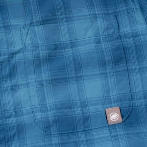 Puede incluir: Una camisa de cuadros azul con un bolsillo en el pecho que incluye una peque&ntilde;a etiqueta rectangular con el logotipo de un mamut blanco. La tela presenta un sutil patr&oacute;n de cuadr&iacute;cula en varios tonos de azul y un bot&oacute;n azul a juego a la izquierda.