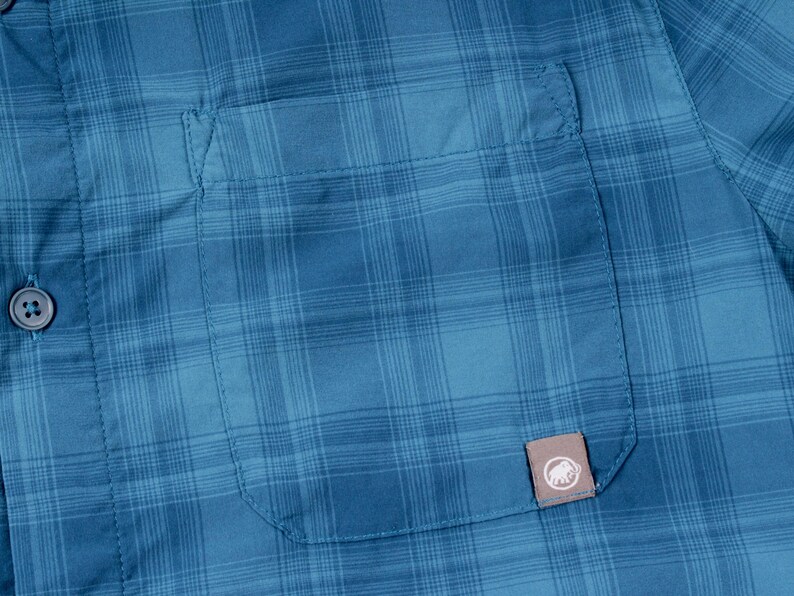 Puede incluir: Una camisa de cuadros azul con un bolsillo en el pecho que incluye una peque&ntilde;a etiqueta rectangular con el logotipo de un mamut blanco. La tela presenta un sutil patr&oacute;n de cuadr&iacute;cula en varios tonos de azul y un bot&oacute;n azul a juego a la izquierda.