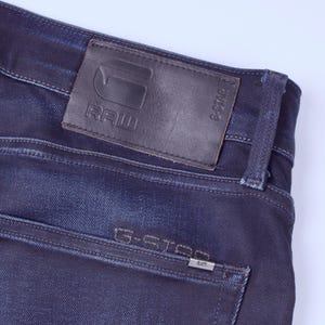 May include: Close-up of dark blue denim jeans. A brown leather patch with the embossed text "RAW" and the G-Star logo is visible. The back pocket features the text "G-STAR" and a small metal tag.
