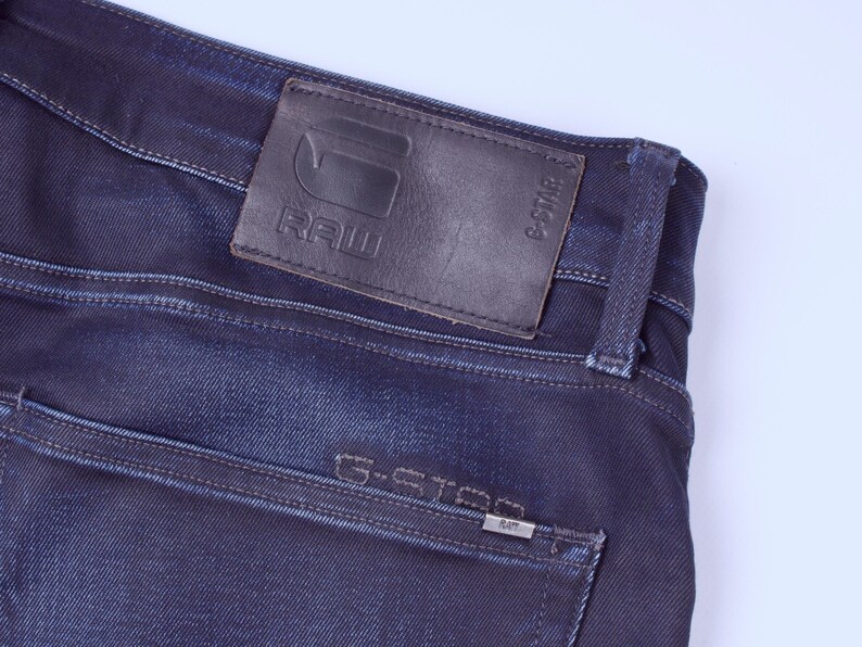 May include: Close-up of dark blue denim jeans. A brown leather patch with the embossed text "RAW" and the G-Star logo is visible. The back pocket features the text "G-STAR" and a small metal tag.
