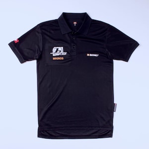 Puede incluir: Polo negro con cuello y mangas cortas. La camiseta presenta los logotipos "HELLVETIA" y "MIGROS" en blanco y naranja, y "BIONIC" en blanco. Un pequeño emblema rojo está en la manga izquierda.