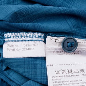 Puede incluir: Una camisa de cuadros azul cerceta con un bot&oacute;n azul oscuro y dos etiquetas de tela blanca. Las etiquetas muestran el texto Style no. 1015-00071, Serial No. 2214088, 831263-7-1-6-ARE-01 P1, Trovat Trail Shirt e instrucciones de cuidado y composici&oacute;n.