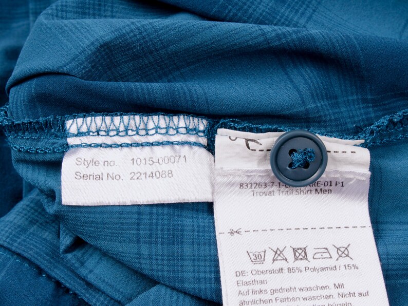 Puede incluir: Una camisa de cuadros azul cerceta con un bot&oacute;n azul oscuro y dos etiquetas de tela blanca. Las etiquetas muestran el texto Style no. 1015-00071, Serial No. 2214088, 831263-7-1-6-ARE-01 P1, Trovat Trail Shirt e instrucciones de cuidado y composici&oacute;n.