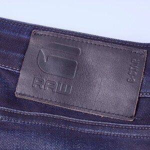 May include: Close-up of dark blue denim jeans with a black leather patch. The patch features the embossed text "RAW" and the G-Star logo. The jeans have visible stitching and a classic denim texture.