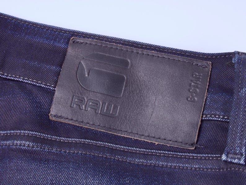 May include: Close-up of dark blue denim jeans with a black leather patch. The patch features the embossed text "RAW" and the G-Star logo. The jeans have visible stitching and a classic denim texture.