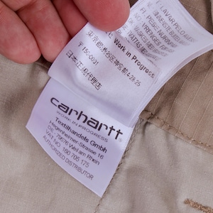 Pode incluir: Close-up de uma etiqueta Carhartt Work In Progress, segurada por uma m&atilde;o, contra um fundo de tecido bege. A etiqueta inclui o logotipo da empresa, o endere&ccedil;o da Textilhandels Gmbh e texto em v&aacute;rios idiomas.