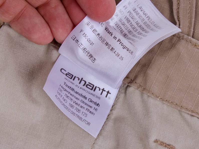 Pode incluir: Close-up de uma etiqueta Carhartt Work In Progress, segurada por uma m&atilde;o, contra um fundo de tecido bege. A etiqueta inclui o logotipo da empresa, o endere&ccedil;o da Textilhandels Gmbh e texto em v&aacute;rios idiomas.