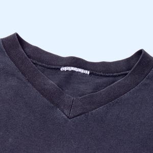 May include: Close-up of a dark gray V-neck t-shirt. The neckline is a darker shade of gray, with a white tag visible at the back of the neck. The fabric appears soft and slightly worn.