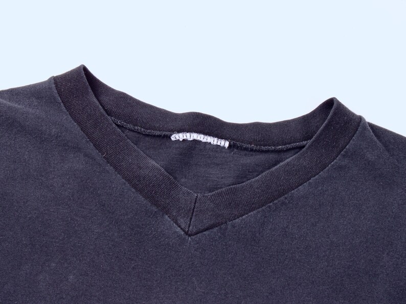 May include: Close-up of a dark gray V-neck t-shirt. The neckline is a darker shade of gray, with a white tag visible at the back of the neck. The fabric appears soft and slightly worn.