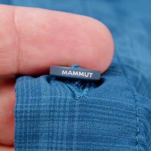Puede incluir: Un bot&oacute;n azul con textura de cuadr&iacute;cula sobre una prenda de tela azul. El bot&oacute;n es rectangular, de color gris oscuro, y presenta la palabra MAMMUT impresa en letras may&uacute;sculas blancas en su borde superior. El bot&oacute;n est&aacute; sujeto con hilo azul.