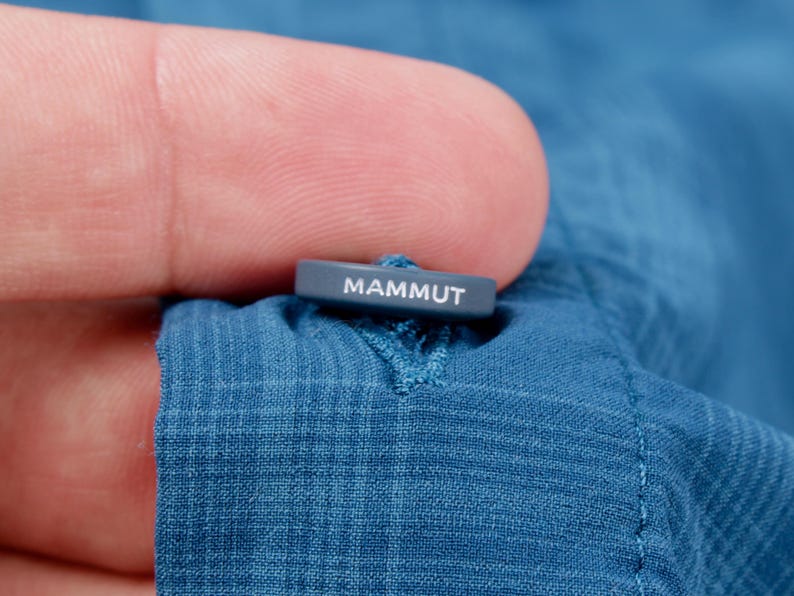 Puede incluir: Un bot&oacute;n azul con textura de cuadr&iacute;cula sobre una prenda de tela azul. El bot&oacute;n es rectangular, de color gris oscuro, y presenta la palabra MAMMUT impresa en letras may&uacute;sculas blancas en su borde superior. El bot&oacute;n est&aacute; sujeto con hilo azul.