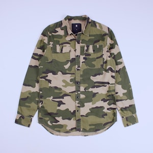 Op de afbeelding: Een shirt met lange mouwen en een camouflagepatroon in groen, bruin en beige. Het shirt heeft een knoopsluiting, twee borstzakken en een kraag. De stof lijkt slijtvast en geschikt voor dagelijks gebruik.