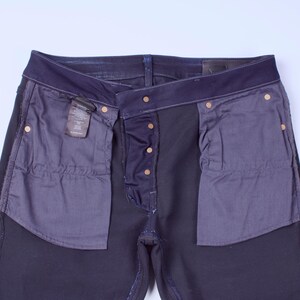 May include: Dark blue denim jeans with a button fly and brass-colored rivets. The pockets are lined with a contrasting dark gray fabric. A small tag is visible near the front pocket.