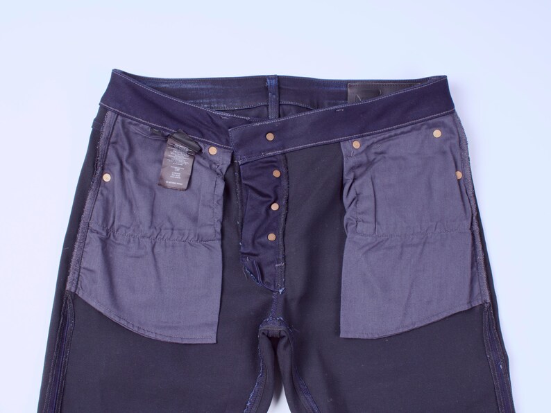 May include: Dark blue denim jeans with a button fly and brass-colored rivets. The pockets are lined with a contrasting dark gray fabric. A small tag is visible near the front pocket.