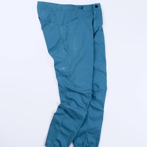 May include: A pair of teal blue trousers featuring a button closure at the waist, side pockets, and a small embroidered logo on the upper thigh area. The pants have a straight-leg cut with visible seam detailing along the sides and front.