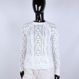May include: A white, long-sleeved sweater with a crew neck and open knit design. The sweater features a detailed pattern of vertical lines and small openings, creating a textured look. The garment is displayed on a mannequin.