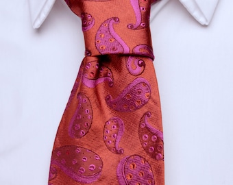 May include: A close-up of a necktie with a paisley pattern. The tie is a burnt orange color with a repeating pattern of pink paisley shapes. The tie is knotted and worn with a white collared shirt.