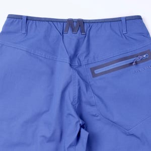 Puede incluir: Primer plano de la parte trasera de unos pantalones azules para actividades al aire libre. Cuenta con un bolsillo con cremallera con la marca "MAMMUT" y un distintivo logotipo "M" en la cintura. Los pantalones tienen un corte a medida y costuras visibles.