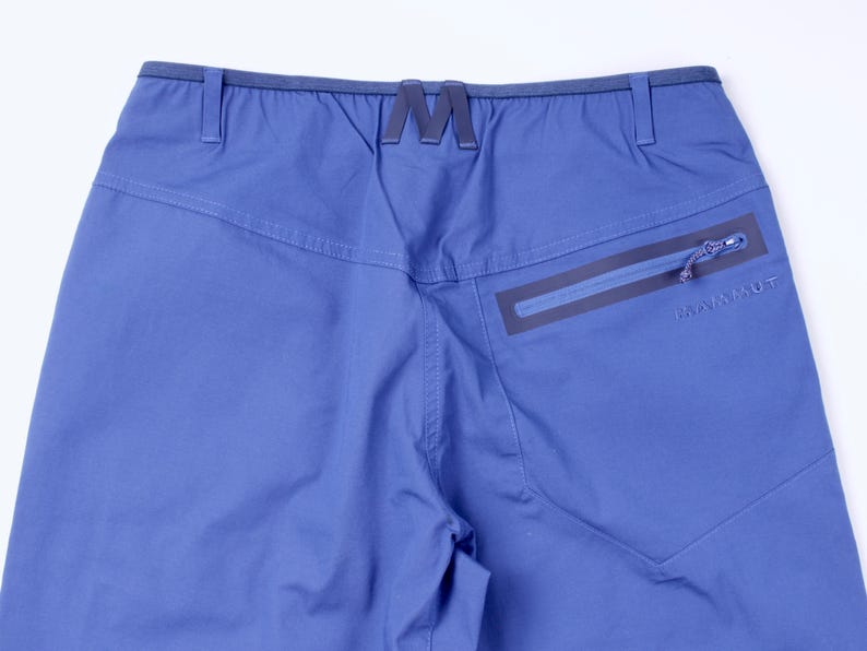 Puede incluir: Primer plano de la parte trasera de unos pantalones azules para actividades al aire libre. Cuenta con un bolsillo con cremallera con la marca "MAMMUT" y un distintivo logotipo "M" en la cintura. Los pantalones tienen un corte a medida y costuras visibles.