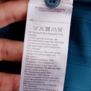 Puede incluir: Una etiqueta de cuidado blanca unida a una camisa de cuadros azul cerceta con un bot&oacute;n azul. La etiqueta muestra el texto 831263-7-1-LAB CARE-01 P1 Trovat Trail Shirt, s&iacute;mbolos de lavado y una composici&oacute;n de 85% poliamida y 15% elastano.