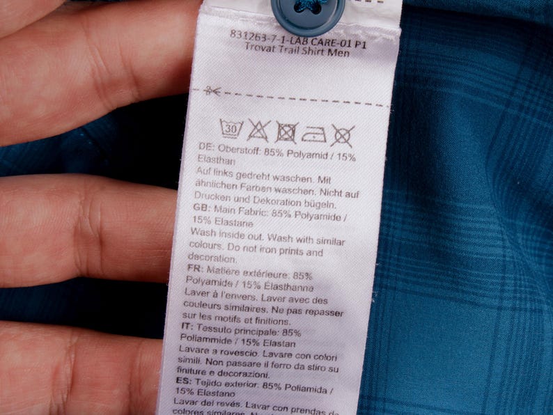 Puede incluir: Una etiqueta de cuidado blanca unida a una camisa de cuadros azul cerceta con un bot&oacute;n azul. La etiqueta muestra el texto 831263-7-1-LAB CARE-01 P1 Trovat Trail Shirt, s&iacute;mbolos de lavado y una composici&oacute;n de 85% poliamida y 15% elastano.