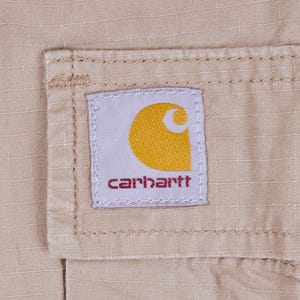 Pode incluir: Close-up de um tecido bege com uma etiqueta costurada. A etiqueta &eacute; branca com uma meia-lua amarela e a palavra "carhartt" em vermelho. O tecido tem um padr&atilde;o de grade subtil.