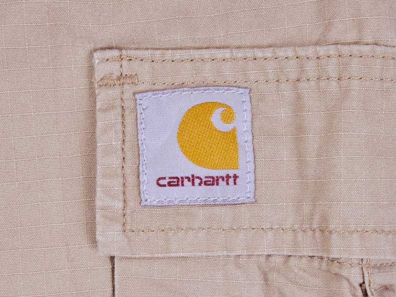 Pode incluir: Close-up de um tecido bege com uma etiqueta costurada. A etiqueta &eacute; branca com uma meia-lua amarela e a palavra "carhartt" em vermelho. O tecido tem um padr&atilde;o de grade subtil.