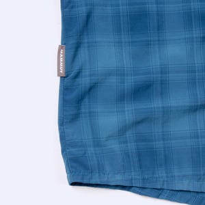 Puede incluir: Una camisa de cuadros azul con un sutil patr&oacute;n de rejilla en varios tonos de azul. Una peque&ntilde;a etiqueta de tela gris rectangular est&aacute; cosida en la costura lateral, mostrando el nombre de la marca MAMMUT en letras may&uacute;sculas blancas.