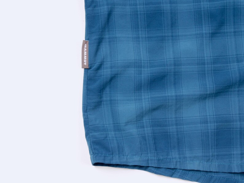 Puede incluir: Una camisa de cuadros azul con un sutil patr&oacute;n de rejilla en varios tonos de azul. Una peque&ntilde;a etiqueta de tela gris rectangular est&aacute; cosida en la costura lateral, mostrando el nombre de la marca MAMMUT en letras may&uacute;sculas blancas.