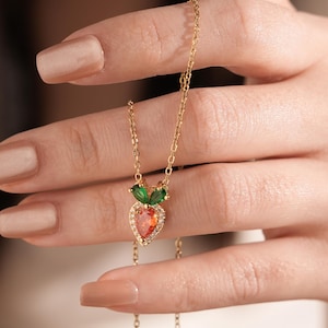 May include: A gold chain necklace with a pendant shaped like a strawberry. The strawberry is made of red and clear stones and has two green leaves.