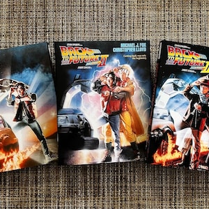 May include: Three movie poster covers for the Back to the Future trilogy. The first poster features a DeLorean time machine with flames coming from the back wheels. The second poster features Michael J. Fox and Christopher Lloyd standing in front of a DeLorean. The third poster features Michael J. Fox, Christopher Lloyd, and Mary Steenburgen standing in front of a DeLorean.