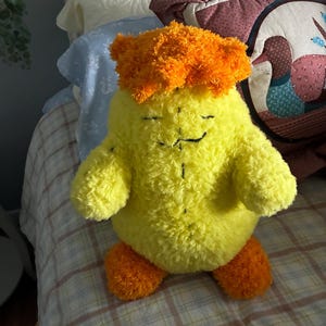 May include: A plush toy shaped like a rounded creature with bright yellow fuzzy fur, orange fuzzy hair on top, and orange fuzzy feet. The face features simple black embroidered lines for closed eyes and a small mouth, with a vertical stitch down the center.