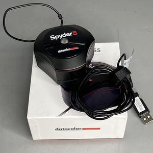 May include: A black Spyder5 colour calibration device with a black cable and USB connector, resting on a white box. The device has the text "Spyder5" and "datacolor" printed on it. The box also has the "datacolor" logo.
