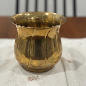 May include: A small, polished brass vessel with a rounded, bulbous body and a flared, scalloped rim. The exterior surface features a series of vertical, swirling indentations that create a textured, reflective appearance across the metallic gold finish.
