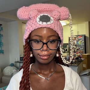 May include: A light pink crocheted hat shaped like a vintage rotary telephone. The hat features a circular grey dial with white numbers and a white coiled cord hanging down the side. The design mimics the handset and base of a classic phone.