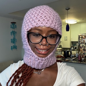Pode incluir: Uma balaclava de crochê roxa clara com um padrão de ponto texturizado. A touca apresenta uma abertura para o rosto e um aquecedor de pescoço. A balaclava é feita de um fio macio e grosso.